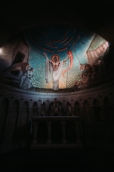 A mosaic depicting a religious scene with a central figure in a white robe, arms raised, haloed with light. Surrounding this central figure are other figures engaged in various postures, suggesting an interaction or worship. The setting appears to be within an arched alcove, with detailed architectural elements including columns and an altar below. The overall ambiance is dimly lit, enhancing the solemn and sacred atmosphere.