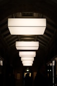 A series of large, rectangular light fixtures hang in a symmetrical arrangement along a hallway with an arched ceiling. The soft, diffused lighting creates a contrast with the dark surroundings, casting shadows on the walls. Silhouetted figures can be seen walking through the corridor, adding a sense of mystery and movement to the scene.