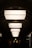 A series of large, rectangular light fixtures hang in a symmetrical arrangement along a hallway with an arched ceiling. The soft, diffused lighting creates a contrast with the dark surroundings, casting shadows on the walls. Silhouetted figures can be seen walking through the corridor, adding a sense of mystery and movement to the scene.
