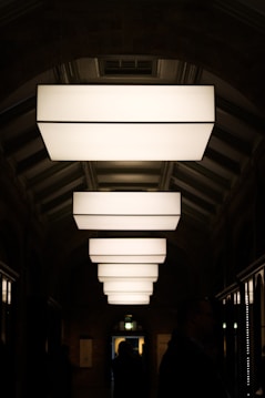A series of large, rectangular light fixtures hang in a symmetrical arrangement along a hallway with an arched ceiling. The soft, diffused lighting creates a contrast with the dark surroundings, casting shadows on the walls. Silhouetted figures can be seen walking through the corridor, adding a sense of mystery and movement to the scene.