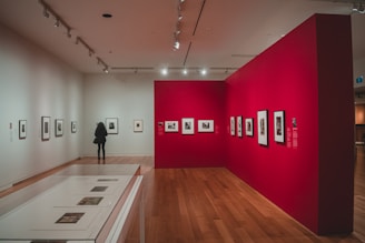A spacious art gallery with wooden floors and bright lighting, featuring walls adorned with framed photographs. The red accent wall contrasts with the neutral tones of the other walls. A person is observing one of the artworks closely.