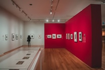 A spacious art gallery with wooden floors and bright lighting, featuring walls adorned with framed photographs. The red accent wall contrasts with the neutral tones of the other walls. A person is observing one of the artworks closely.