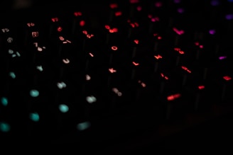 A close-up view of a keyboard with backlit keys. The keys are illuminated in various colors including red, blue, and green, creating a vibrant and visually striking effect. The keys are in focus with some blurring in the background, adding to the depth of the image.