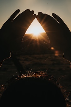 Hands are forming a shape resembling a triangle or heart in front of a bright sun. The sun is positioned at the center, casting a warm glow that creates a silhouette effect. Hair strands are visible at the bottom, catching light from the sun.