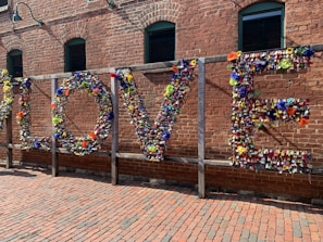 A large installation on a brick wall spelling out the word 'LOVE' made entirely of colorful padlocks and flowers. The background consists of an industrial-style red brick building with green-framed windows. Sunlight casts soft shadows on the ground, which is paved with bricks, creating a warm atmosphere.