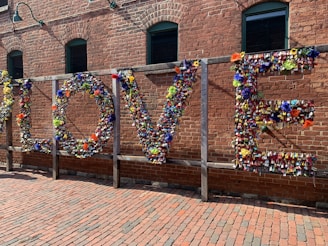 A large installation on a brick wall spelling out the word 'LOVE' made entirely of colorful padlocks and flowers. The background consists of an industrial-style red brick building with green-framed windows. Sunlight casts soft shadows on the ground, which is paved with bricks, creating a warm atmosphere.