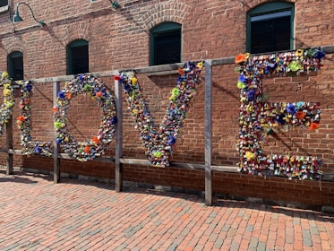 A large installation on a brick wall spelling out the word 'LOVE' made entirely of colorful padlocks and flowers. The background consists of an industrial-style red brick building with green-framed windows. Sunlight casts soft shadows on the ground, which is paved with bricks, creating a warm atmosphere.