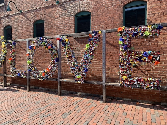 A large installation on a brick wall spelling out the word 'LOVE' made entirely of colorful padlocks and flowers. The background consists of an industrial-style red brick building with green-framed windows. Sunlight casts soft shadows on the ground, which is paved with bricks, creating a warm atmosphere.