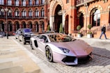 A sleek, silver luxury sports car is parked on a cobblestone street in front of a historic red brick building with detailed arches. Another luxury vehicle is parked behind it. The scene exudes elegance and urban sophistication, with a suited man walking nearby.