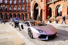 A sleek, silver luxury sports car is parked on a cobblestone street in front of a historic red brick building with detailed arches. Another luxury vehicle is parked behind it. The scene exudes elegance and urban sophistication, with a suited man walking nearby.