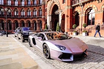 A sleek, silver luxury sports car is parked on a cobblestone street in front of a historic red brick building with detailed arches. Another luxury vehicle is parked behind it. The scene exudes elegance and urban sophistication, with a suited man walking nearby.
