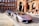 A sleek, silver luxury sports car is parked on a cobblestone street in front of a historic red brick building with detailed arches. Another luxury vehicle is parked behind it. The scene exudes elegance and urban sophistication, with a suited man walking nearby.