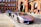 A sleek, silver luxury sports car is parked on a cobblestone street in front of a historic red brick building with detailed arches. Another luxury vehicle is parked behind it. The scene exudes elegance and urban sophistication, with a suited man walking nearby.