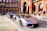 A sleek, silver luxury sports car is parked on a cobblestone street in front of a historic red brick building with detailed arches. Another luxury vehicle is parked behind it. The scene exudes elegance and urban sophistication, with a suited man walking nearby.