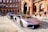 A sleek, silver luxury sports car is parked on a cobblestone street in front of a historic red brick building with detailed arches. Another luxury vehicle is parked behind it. The scene exudes elegance and urban sophistication, with a suited man walking nearby.