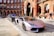 A sleek, silver luxury sports car is parked on a cobblestone street in front of a historic red brick building with detailed arches. Another luxury vehicle is parked behind it. The scene exudes elegance and urban sophistication, with a suited man walking nearby.