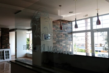 The interior of a modern office space with large windows that let in natural light. The walls are partially covered with decorative stone. There are hanging light fixtures with red shades, and the reflection of a logo, possibly an eagle, can be seen on the glass partition. Outside, there are palm trees and a view of the street.