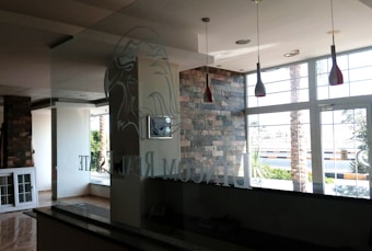 The interior of a modern office space with large windows that let in natural light. The walls are partially covered with decorative stone. There are hanging light fixtures with red shades, and the reflection of a logo, possibly an eagle, can be seen on the glass partition. Outside, there are palm trees and a view of the street.