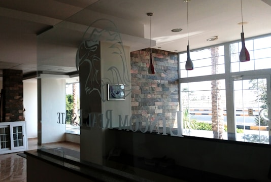 The interior of a modern office space with large windows that let in natural light. The walls are partially covered with decorative stone. There are hanging light fixtures with red shades, and the reflection of a logo, possibly an eagle, can be seen on the glass partition. Outside, there are palm trees and a view of the street.