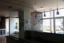 The interior of a modern office space with large windows that let in natural light. The walls are partially covered with decorative stone. There are hanging light fixtures with red shades, and the reflection of a logo, possibly an eagle, can be seen on the glass partition. Outside, there are palm trees and a view of the street.