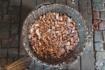 A woven basket filled with wood shavings sits on a rough, wooden and cobblestone floor. The shavings are light brown and curly, indicating they are likely from a woodworking project. A broom with natural bristles rests beside the basket.