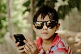 A young child wearing dark sunglasses and a multicolored striped shirt is holding a smartphone in their hands. The background consists of blurred greenery possibly surrounded by a chicken wire fence, creating a natural and relaxed setting.