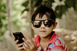 A young child wearing dark sunglasses and a multicolored striped shirt is holding a smartphone in their hands. The background consists of blurred greenery possibly surrounded by a chicken wire fence, creating a natural and relaxed setting.