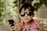 A young child wearing dark sunglasses and a multicolored striped shirt is holding a smartphone in their hands. The background consists of blurred greenery possibly surrounded by a chicken wire fence, creating a natural and relaxed setting.