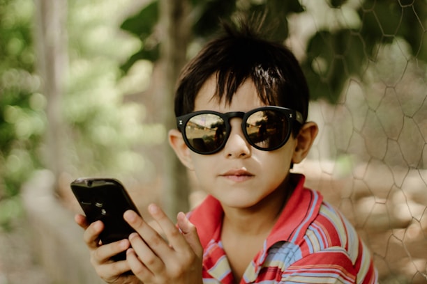 A young child wearing dark sunglasses and a multicolored striped shirt is holding a smartphone in their hands. The background consists of blurred greenery possibly surrounded by a chicken wire fence, creating a natural and relaxed setting.