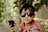 A young child wearing dark sunglasses and a multicolored striped shirt is holding a smartphone in their hands. The background consists of blurred greenery possibly surrounded by a chicken wire fence, creating a natural and relaxed setting.