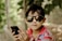 A young child wearing dark sunglasses and a multicolored striped shirt is holding a smartphone in their hands. The background consists of blurred greenery possibly surrounded by a chicken wire fence, creating a natural and relaxed setting.