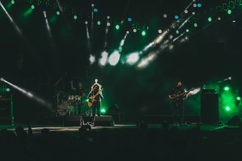 A live music performance on stage features three musicians. Vibrant green and white stage lights create a dramatic atmosphere. A drummer is visible in the background, while two guitarists stand at the front, illuminated by spotlights. Fog or smoke enhances the visual effect, contributing to the dynamic mood.