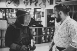 Two individuals are engaged in a conversation in what appears to be a martial arts dojo. One person is wearing traditional kendo gear, including a helmet and armor, while holding a shinai, a bamboo sword. The other individual is dressed in a patterned robe typical of martial arts attire. The background shows wooden shelves holding various objects.