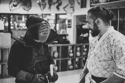 Two individuals are engaged in a conversation in what appears to be a martial arts dojo. One person is wearing traditional kendo gear, including a helmet and armor, while holding a shinai, a bamboo sword. The other individual is dressed in a patterned robe typical of martial arts attire. The background shows wooden shelves holding various objects.
