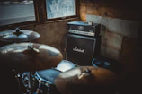 Vintage amplifier and drum set beside a crate of Ritmo Ámbar beers in a gritty rehearsal space.