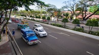 A busy urban street with multiple vehicles, including colorful jeepneys, cars, and motorcycles. Lush green trees and well-maintained hedges line the street, with buildings in the background. Pedestrians can be seen on the sidewalk, and the atmosphere is lively with ample daylight.