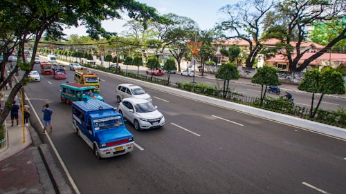 A busy urban street with multiple vehicles, including colorful jeepneys, cars, and motorcycles. Lush green trees and well-maintained hedges line the street, with buildings in the background. Pedestrians can be seen on the sidewalk, and the atmosphere is lively with ample daylight.