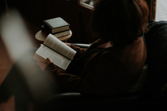 A person is sitting in a dimly lit room, reading a book with pages open. There are additional books stacked on a small round table nearby. The setting suggests a cozy reading nook with subdued lighting and a relaxed atmosphere.