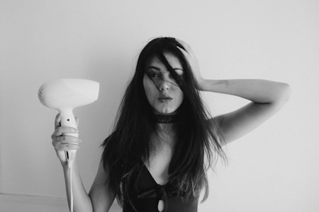 A woman stands against a plain background holding a hair dryer. Her long hair partially covers her face, and she has one hand placed on her head. The expression appears serious or contemplative.