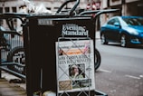 A newspaper stand on a city street displaying the London Evening Standard with a headline about the coronavirus. The stand has a black metal frame with a sign that reads 'FREE PLEASE TAKE ONE'. There is a blue car parked on the street in the background, along with blurred cityscape elements.
