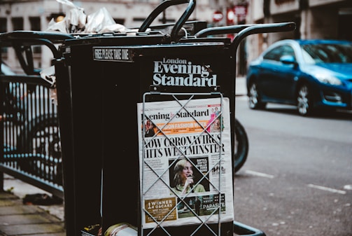 A newspaper stand on a city street displaying the London Evening Standard with a headline about the coronavirus. The stand has a black metal frame with a sign that reads 'FREE PLEASE TAKE ONE'. There is a blue car parked on the street in the background, along with blurred cityscape elements.