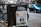 A newspaper stand on a city street displaying the London Evening Standard with a headline about the coronavirus. The stand has a black metal frame with a sign that reads 'FREE PLEASE TAKE ONE'. There is a blue car parked on the street in the background, along with blurred cityscape elements.
