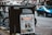 A newspaper stand on a city street displaying the London Evening Standard with a headline about the coronavirus. The stand has a black metal frame with a sign that reads 'FREE PLEASE TAKE ONE'. There is a blue car parked on the street in the background, along with blurred cityscape elements.