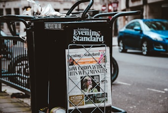 A newspaper stand on a city street displaying the London Evening Standard with a headline about the coronavirus. The stand has a black metal frame with a sign that reads 'FREE PLEASE TAKE ONE'. There is a blue car parked on the street in the background, along with blurred cityscape elements.