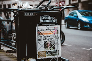 A newspaper stand on a city street displaying the London Evening Standard with a headline about the coronavirus. The stand has a black metal frame with a sign that reads 'FREE PLEASE TAKE ONE'. There is a blue car parked on the street in the background, along with blurred cityscape elements.