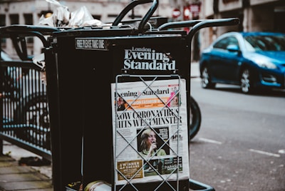 A newspaper stand on a city street displaying the London Evening Standard with a headline about the coronavirus. The stand has a black metal frame with a sign that reads 'FREE PLEASE TAKE ONE'. There is a blue car parked on the street in the background, along with blurred cityscape elements.