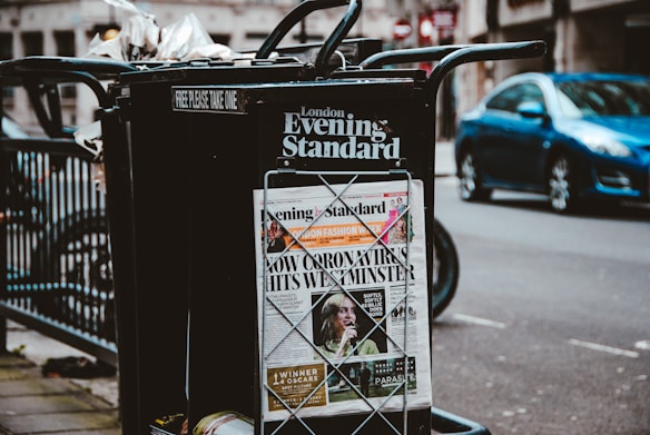 A newspaper stand on a city street displaying the London Evening Standard with a headline about the coronavirus. The stand has a black metal frame with a sign that reads 'FREE PLEASE TAKE ONE'. There is a blue car parked on the street in the background, along with blurred cityscape elements.