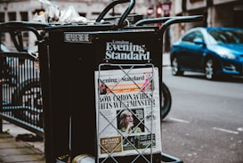 A newspaper stand on a city street displaying the London Evening Standard with a headline about the coronavirus. The stand has a black metal frame with a sign that reads 'FREE PLEASE TAKE ONE'. There is a blue car parked on the street in the background, along with blurred cityscape elements.