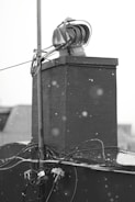 A black and white image depicting a rooftop structure with a ventilation unit and wiring. The background is blurred, and snowflakes are visible in the foreground, indicating a wintery setting.