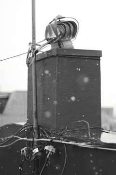 A black and white image depicting a rooftop structure with a ventilation unit and wiring. The background is blurred, and snowflakes are visible in the foreground, indicating a wintery setting.